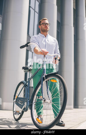 Cool young bearded hipster with bicycle on the city street. Smiling casual businessman having a break. He is standing in front of the building next to Stock Photo