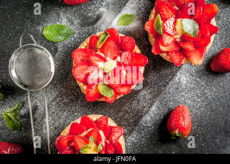 Summer and spring dessert. Home pies tartlets with custard and strawberries, decorated with mint and powdered sugar. On black stone table, rustic, wit Stock Photo