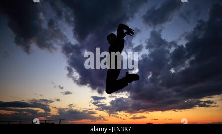 Silhouette of a man against a background of clouds and sunset. He is jumping on the roof. Parkour in the evening. Stock Photo