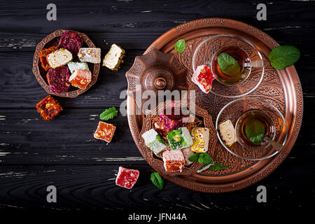 Bowl with various pieces of turkish delight lokum and black tea with mint on a dark wooden background. Flat lay. Top view Stock Photo