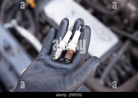 Auto mechanic wearing protective work glove holds old and new spark plugs over a car engine Stock Photo