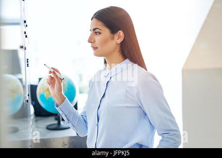 Serious young woman in blue at white board in classroom or small office with globe in background