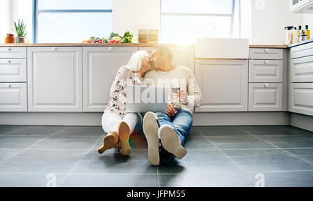Affectionate senior couple sitting on their kitchen floor at home drinking red wine and browsing the internet on a laptop Stock Photo