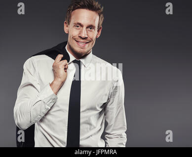 Handsome young businessman smiling and standing alone in a studio against a grey background carrying his jacket over his shoulder Stock Photo