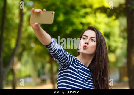 beautiful young brunette caucasian woman taking funny selfie with smartphone outdoors in park. Summer, people, technology, lifestyle concept. Girl mak Stock Photo