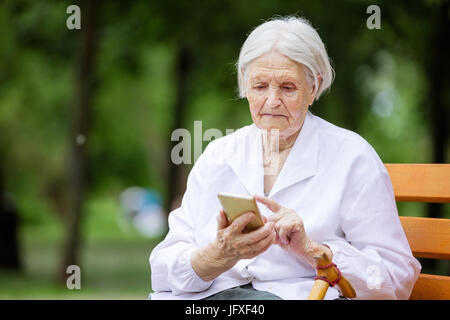 Senior woman using smartphone while sitting on bench in summer park Stock Photo