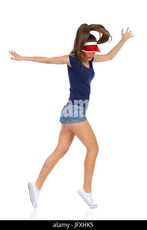 Woman in blue shirt, jeans shorts white sneakers and red sun visor cap dancing tip toe. Full length studio shot isolated on white. Stock Photo