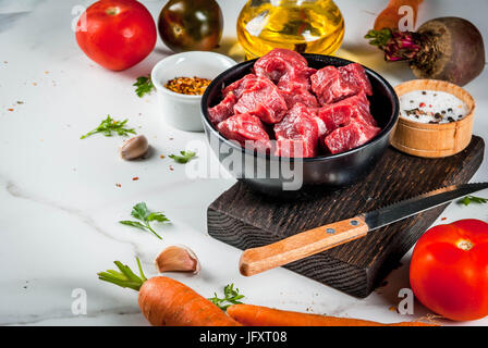 Meat, beef. Fresh raw chopped goulash, beef cubes in a bowl. Spices (salt, pepper), tomatoes, garlic, onions. On a white marble table, with a fork for Stock Photo