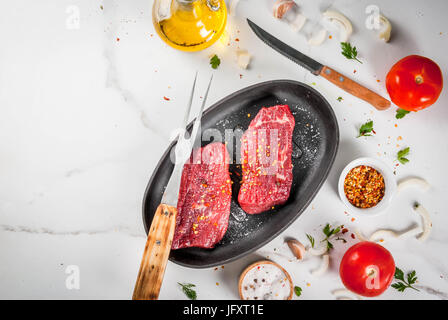 Meat, beef. Fresh raw steaks in a frying pan. Spices (salt, pepper), fresh vegetables - tomatoes, carrots, garlic, onions. On a white marble table, wi Stock Photo
