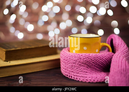 Hot tea, hot chocolate, coffee in yellow cup, wrapped with a pink knitted scarf.  Old books. Blurred lights, wooden background. Winter time, Rustic ba Stock Photo