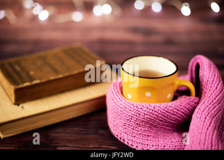 Hot tea, hot chocolate, coffee in yellow cup, wrapped with a pink knitted scarf.  Old books. Blurred lights, wooden background. Winter time, Rustic ba Stock Photo