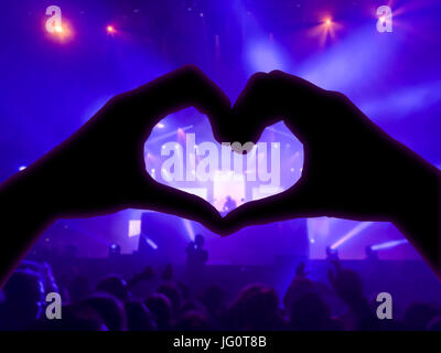 Music concert, hands raised in the shape of the heart for the music, blurred crowd and artists on stage in the background with blue spotlights Stock Photo