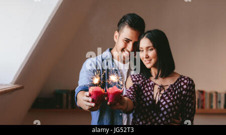 Young couple holding two cupcakes and celebrating their anniversary. Cupcakes with sparklers in hands of couple standing together at home. Stock Photo