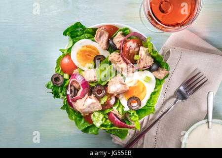 Overhead photo of a plate of salad with canned tuna, boiled eggs, green lettuce leaves, onions, black olives, and cherry tomatoes, on teal texture wit Stock Photo