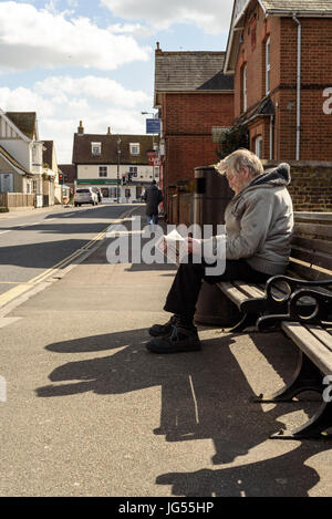 Old man with a beard sits on a bench on the side of a road reading the popular English Sun Newspaper in daylight Stock Photo