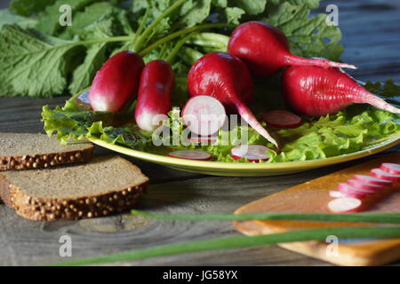 Still life with radish, lettuce, green onions and bread. Radish and lettuce leaves lie on a plate. Clean radishes prepared for use in food. Stock Photo
