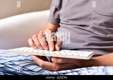 closeup of a young caucasian man in pajamas sitting in a comfortable armchair typing in a computer keyboard Stock Photo