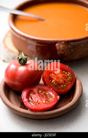 a rustic brown earthenware bowl with tomato soup and some tomatoes in a brown earthenware plate, on a white table Stock Photo