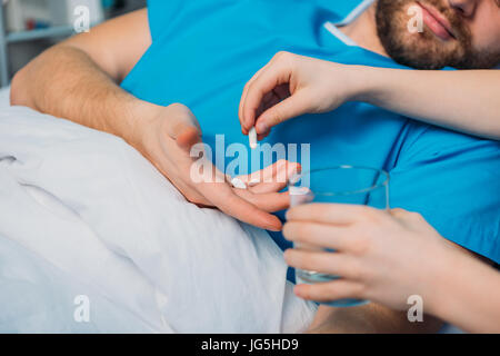 son giving pills and glass of water to his sick father laying at ward, dad and son Stock Photo