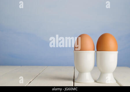 Two fresh brown undecorated eggs, side by side in white porcelain egg cups against a painted blue sky effect background, on an old cream painted wood  Stock Photo
