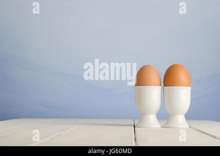 Two fresh brown eggs in egg cups on an old, cream painted wood plank farmhouse table, with painted blue sky effect background. Stock Photo