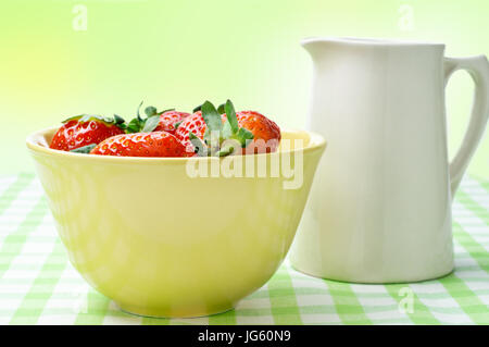 Eye level close up view of a yellow china bowl of red strawberries and a cream jug, on a green and white gingham table cloth with green, yellow and wh Stock Photo