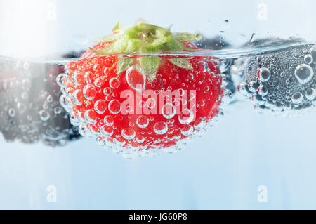 Close up (macro) of a bright red fresh strawberry, floating in sparkling water and covered in oxygen bubbles.  Blackberry and blueberries in soft focu Stock Photo