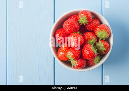 Overhead shot of a white china bowl containing red strawberries with leaves intact, positioned on the right side of a light blue wood planked table. Stock Photo