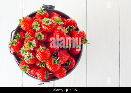 Overhead shot of a tin pail filled to the top with fresh strawberries, on a wooden planked table, painted in soft white.  Copy space to right of frame Stock Photo