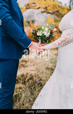 Close-up view of the hands of the newlyweds holding the wedding bouquet of the colourful roses. Stock Photo