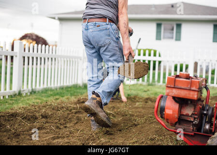 A man shovels dirt in a garden next to a rotatiller. Stock Photo