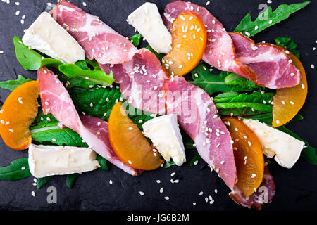 Salad with ham jamon serrano, camembert, melon, arugula on black stone slate plate on black background. Top view. Stock Photo