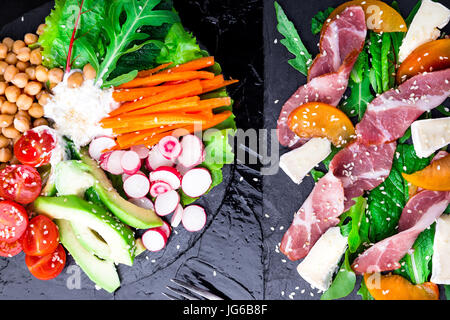 Two different salads om black stone slate. Vegan salad. Meat salad. Salad with ham jamon serrano, camembert, melon, arugula. Top view Stock Photo