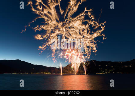 Fireworks on the lakefront of Luino over the Maggiore Lake in a summer evening with blue sky and mountains in the background Stock Photo