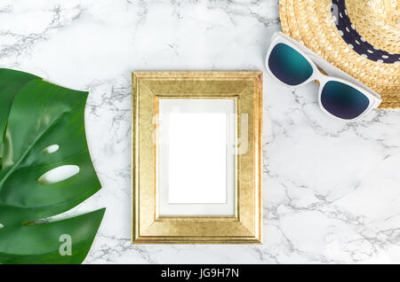 Blank Vintage golden color picture frame on green monstera leaf with sunglasses and straw hat on white marble table top view,Mock up for adding your p Stock Photo