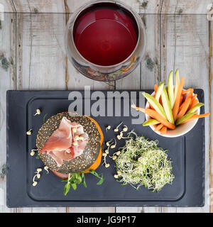 Healthy burger with hamon, tomatoes, micro greens and black wholegrain buns, vegetable sticks and red wine on black slate board over wooden background. Clean eating, dieting, detox food concept. Stock Photo