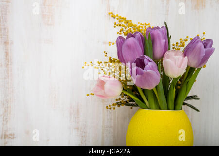 Bouquet of tulips and mimosa in a yellow vase on a wooden background. Still life with beautiful flowers on holiday. Stock Photo