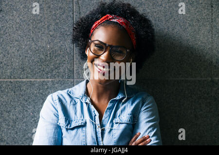 Smiling young African woman wearing glasses and a bandana standing with her arms crossed against a wall listing to music on earphones Stock Photo