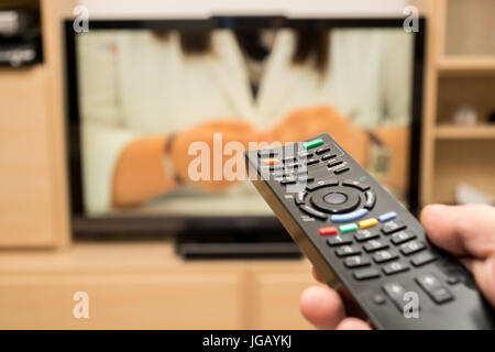 Watching TV and using black modern remote controller. Hand holding TV remote control with a television in the background. Shallow dof. Stock Photo