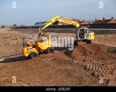 A small yellow tracked crawler excavator working on a beach to lift shingle into a dump truck, UK. Stock Photo