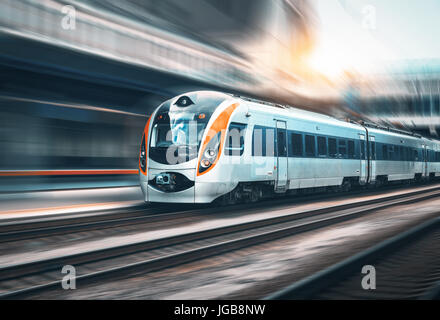 High speed train in motion at the railway station at sunset in Europe. Modern intercity train on the railway platform with motion blur effect. Industr Stock Photo