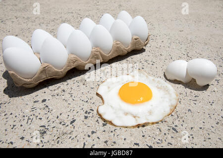 Fried egg with broken shells and carton of white eggs on hot sidewalk on summer day. Stock Photo