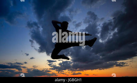 Silhouette of a man against a background of clouds and golden sunset. He is jumping on the roof. Parkour in the evening. Stock Photo