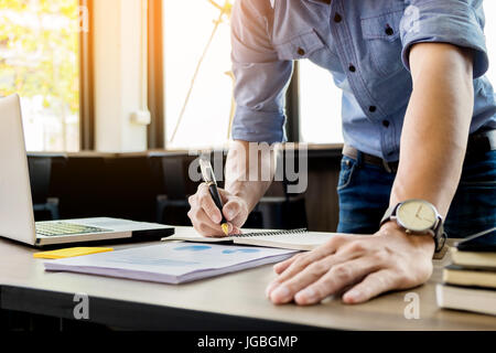 Young business man working in bright office, using laptop, writing notes. Stock Photo