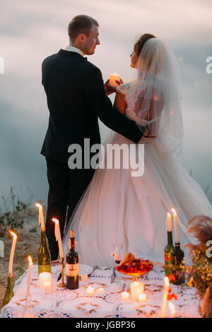 The back view of the newlyweds iwth the shining candle standing behind teh romantic picnic set. Stock Photo