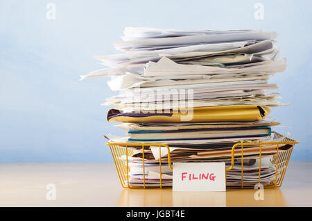 An old yellow wireframe filing tray, piled high with documents and folders, on a light wood veneer desk against light blue background. Stock Photo