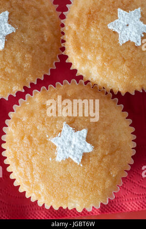 Overhead shot of three vanilla cupcakes in paper cases on red paper napkin, decorated with sprinkled white icing sugar in the shape of stars. Stock Photo