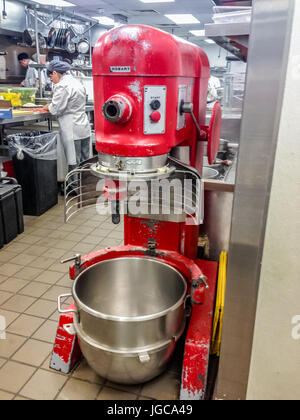Large red industrial mixer with an empty clean stainless steel bowl in a bakery with people working at kitchen counters in the background Stock Photo