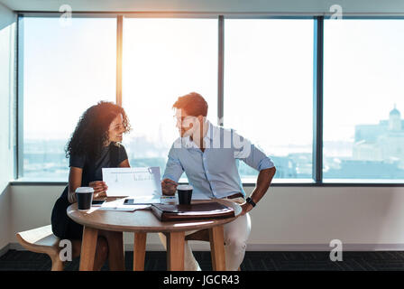 Young man and woman discussing work over a cup of coffee sitting in office. Businesswoman holding a paper in hand while taking to her business colleag Stock Photo
