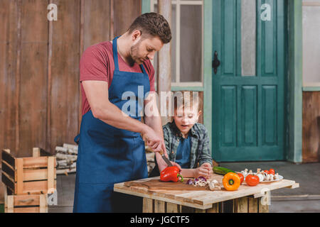 Smiling little boy looking at happy father cutting vegetables on cutting board, dad and son cooking concept Stock Photo